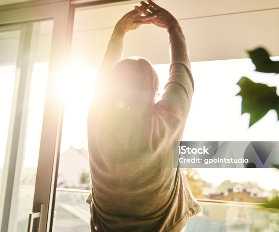 Person mit erhobenen Armen vor einem Fenster, Sonnenstrahlen strahlen durch.