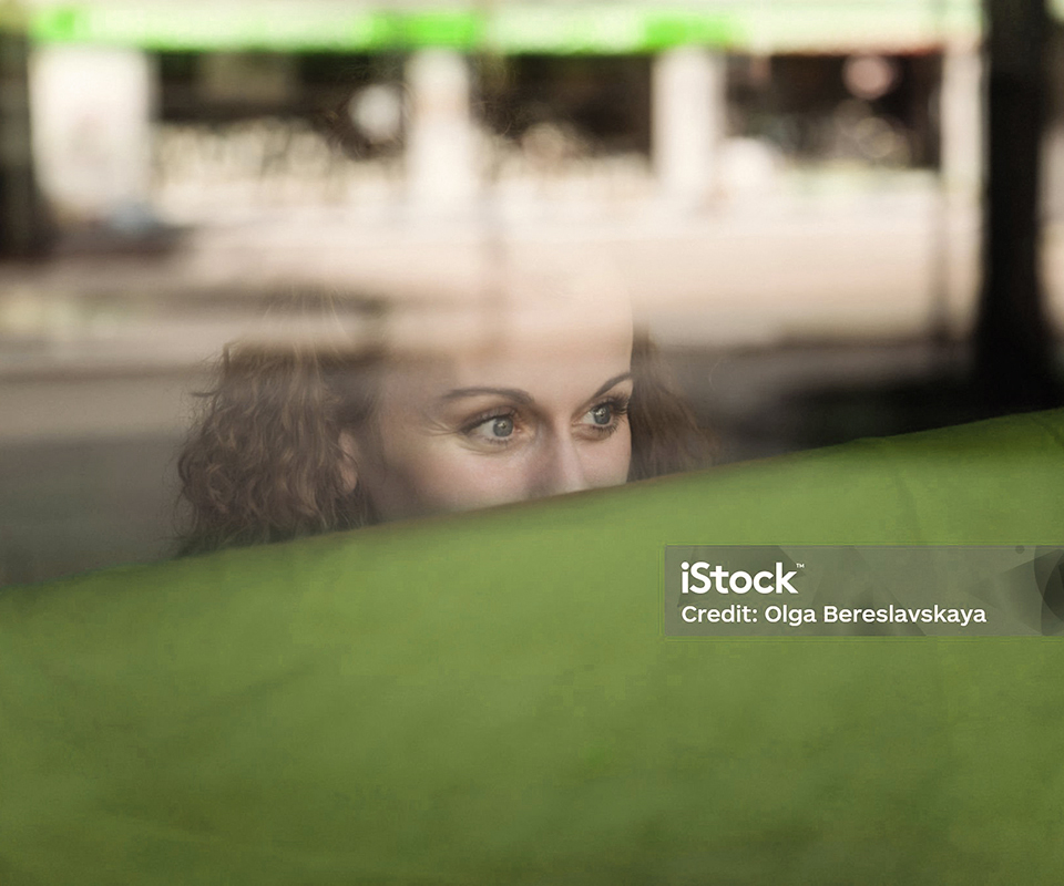 Eine Frau mit Locken blickt neugierig durch ein Fenster, das eine grüne Fläche reflektiert.