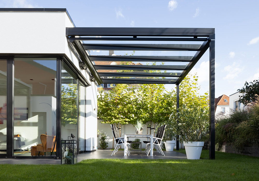 Moderne Überdachung mit Glas und einem Tisch sowie Stühlen in einer Terrassensituation. Dominante Farben: Schwarz und Grün.