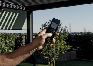 Hand, die eine Fernbedienung hält, mit Blick auf eine Gartenlandschaft und zahlreichen Pflanzen im Hintergrund. 