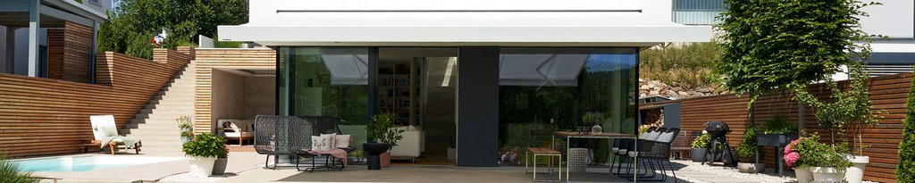 Moderne Terrasse mit Glasfront, Überdachung und Terrassensituation, umgeben von Pflanzen und Holzstrukturen.