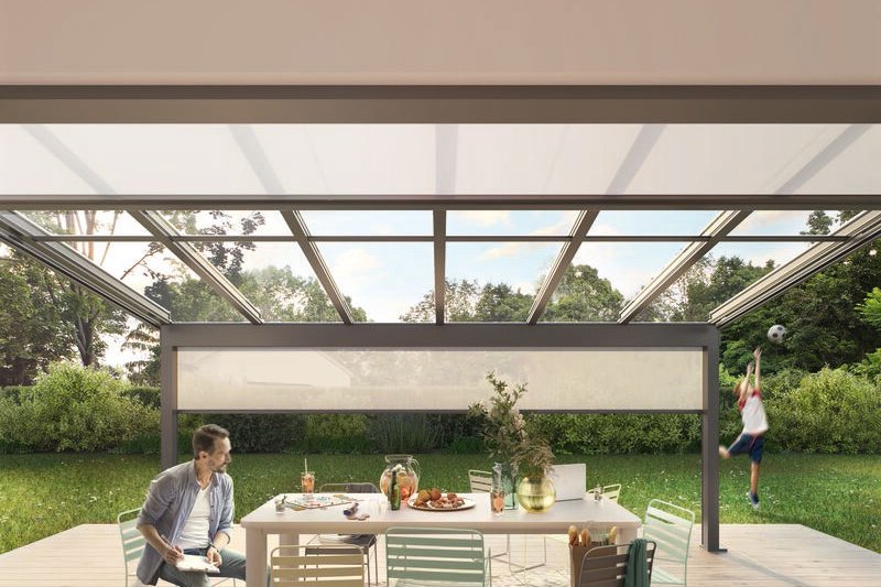 Terrassensituation mit Tisch, Stühlen, Markise und Glas-Schiebewänden, umgeben von Grün und einer Spielszene im Hintergrund. Terrassensituation mit Tisch, Stühlen, Markise und Glas-Schiebewänden, umgeben von Grün und einer Spielszene im Hintergrund.
