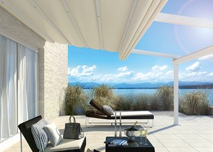 Terrassensituation mit Markise, Überdachung und Blick auf Wasser mit Himmel und Wolken im Hintergrund. 