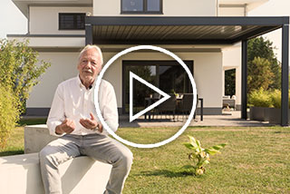 Mann sitzt auf einem Stein, spricht und zeigt auf seine Terrassensituation mit Überdachung im Hintergrund.