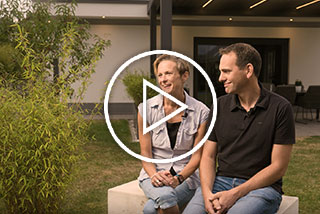 Zwei Personen sitzen auf einer Steinbank in einer Terrassensituation mit Pflanzen im Hintergrund.