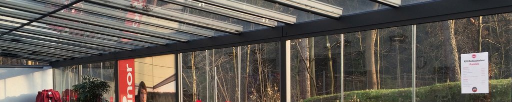 Eine moderne Überdachung mit Glas-Schiebewänden und Sonnenlicht, im Hintergrund ein bewaldeter Bereich.